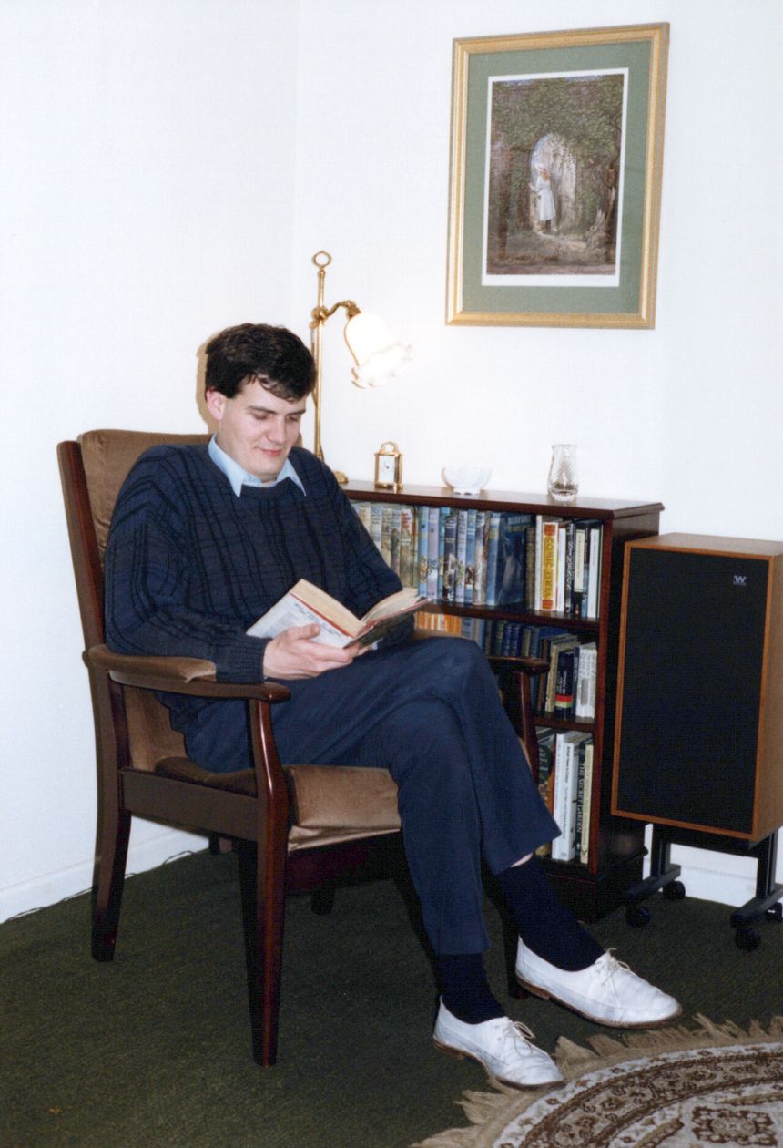 A young Rob in an armchair, reading beside his bookcase